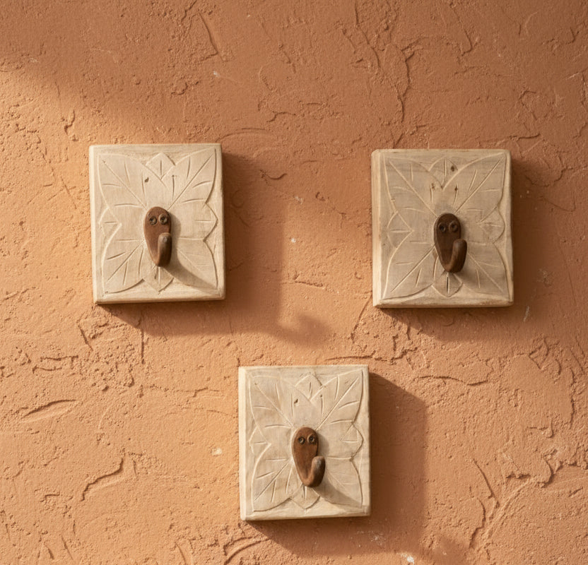 Five square beige stone wall hooks with carved leaf patterns and small brown bird-shaped pendants, arranged on a light cream background.
