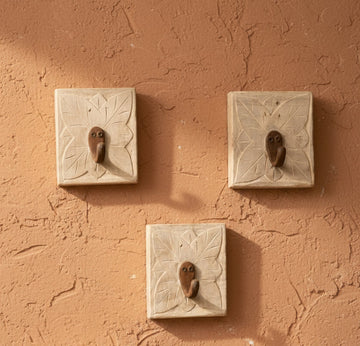 Five square beige stone wall hooks with carved leaf patterns and small brown bird-shaped pendants, arranged on a light cream background.