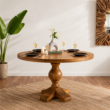 A round wooden dining table with a turned pedestal base, styled with black plates, wine glasses, candles, and a small vase of white flowers in a bright room with a potted plant and woven rug.