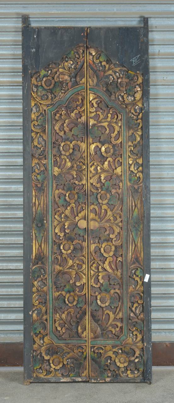 An ornate carved wooden door with intricate floral and scroll patterns in gold, brown, and green tones, set against a corrugated metal background.