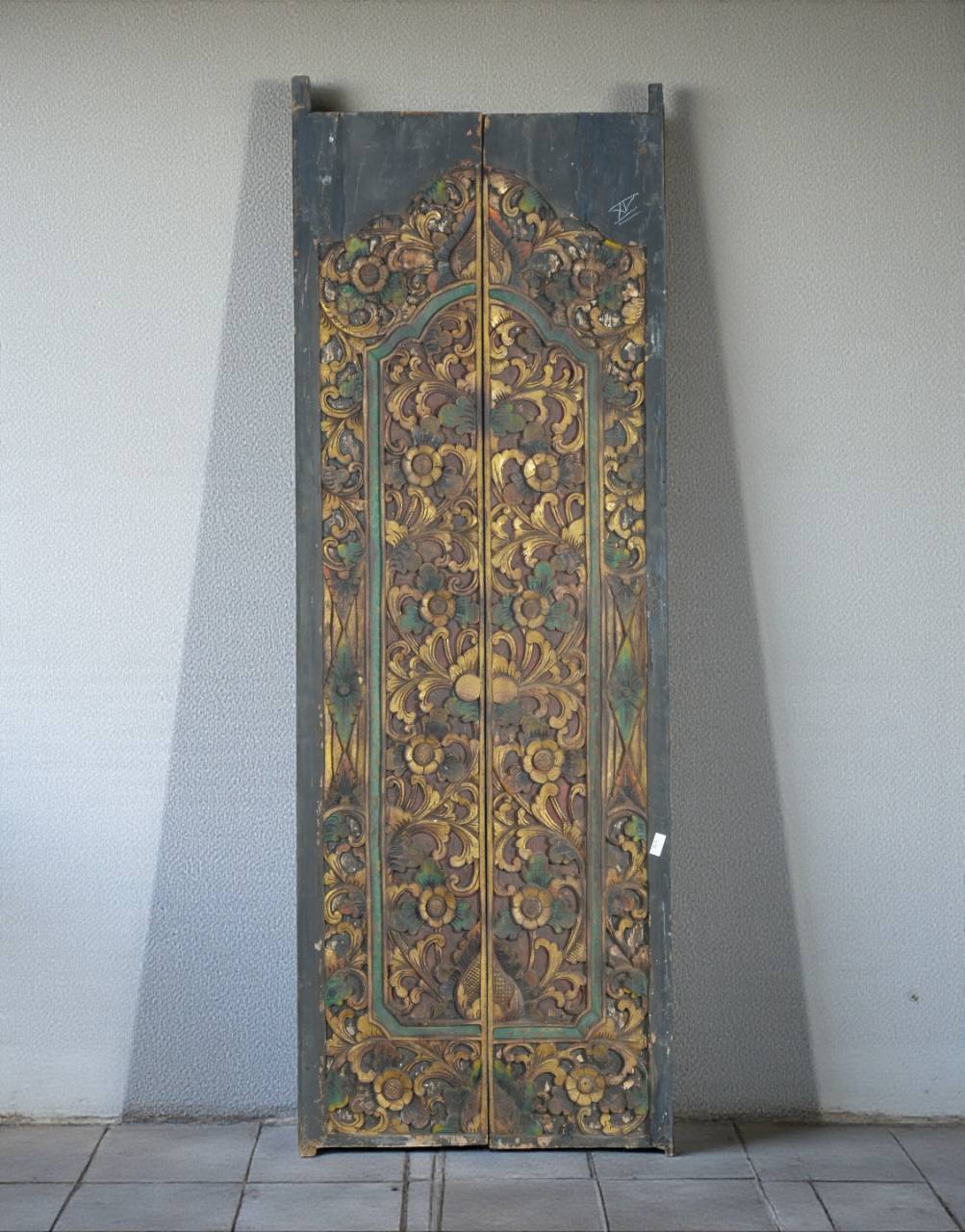 An ornate, tall wooden door panel with intricate hand-painted floral and scrollwork patterns in gold, green, and brown tones, leaning against a plain light gray wall.