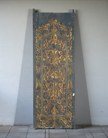 An ornate, tall wooden door panel with intricate hand-painted floral and scrollwork patterns in gold, green, and brown tones, leaning against a plain light gray wall.