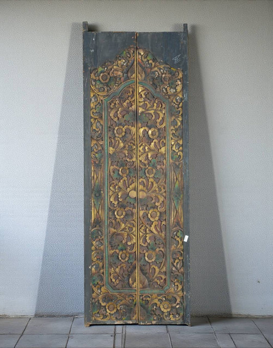 An ornate, tall wooden door panel with intricate hand-painted floral and scrollwork patterns in gold, green, and brown tones, leaning against a plain light gray wall.
