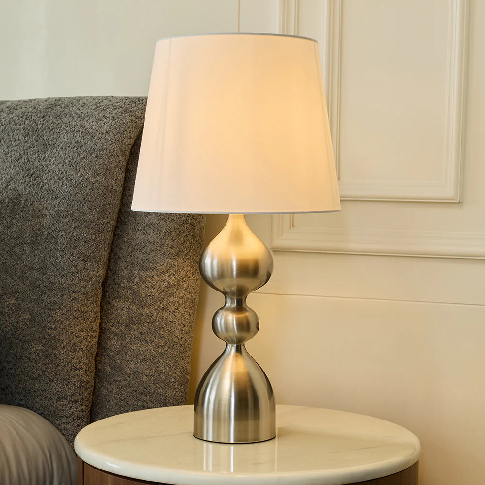 The sleek curves of the brushed metal base add a sophisticated touch, while the neutral fabric shade ensures gentle, diffused lighting that enhances the ambiance of your home.