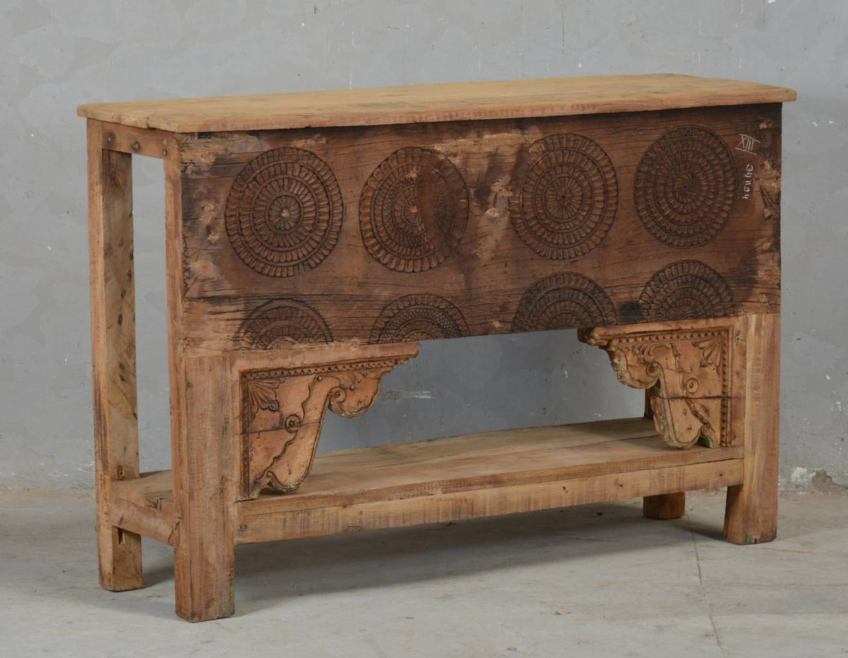 A rustic carved wooden console table with circular patterns on the front panel, a lower shelf, and ornate corner brackets, shown against a plain gray wall.