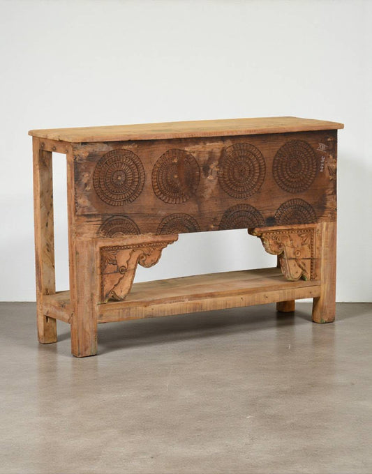 A rustic carved wooden console table with a warm brown finish, featuring circular geometric patterns on the front, decorative corbels, and an open lower shelf, set against a plain light gray wall and concrete floor.