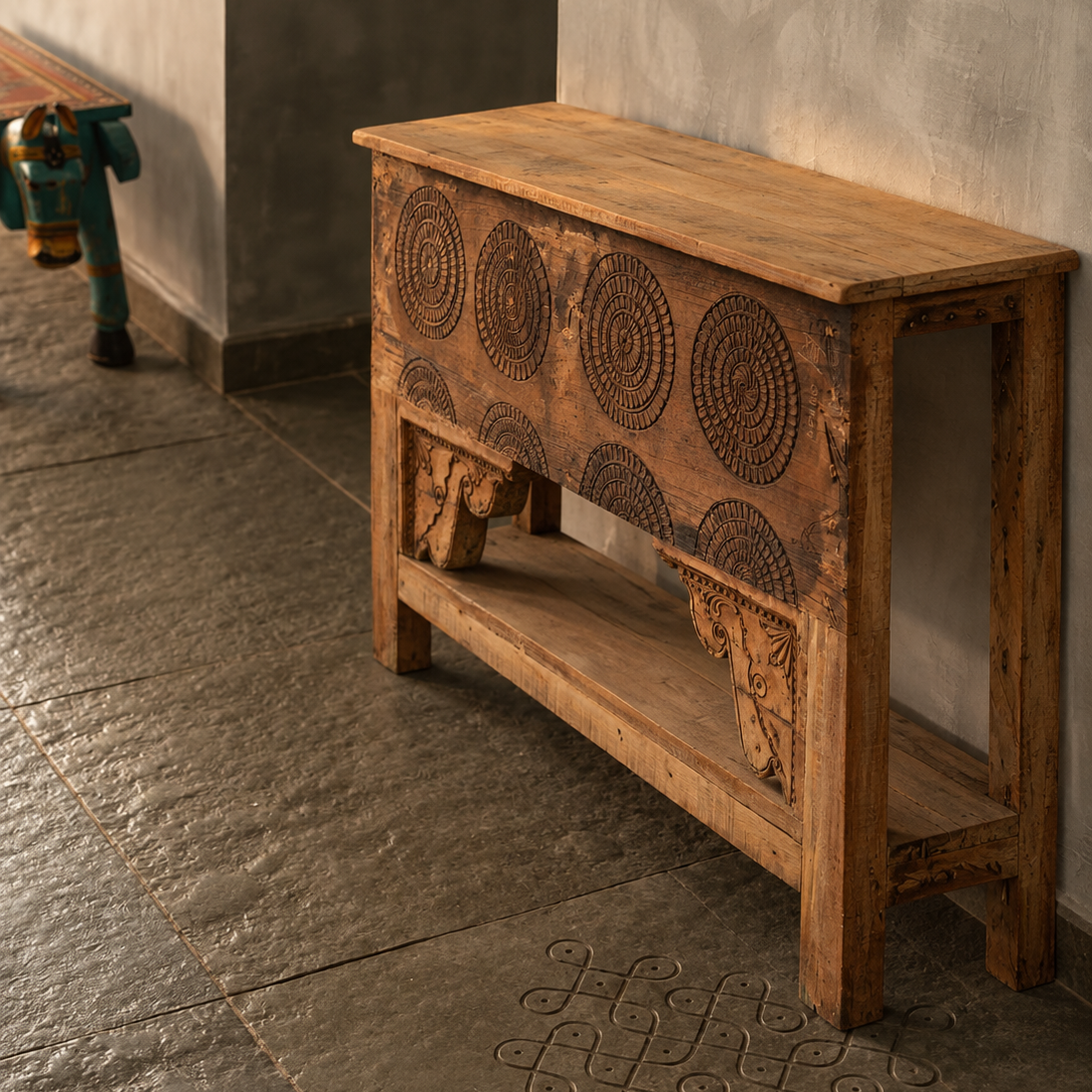 A rustic carved wooden console table with circular geometric engravings on the front, set against a textured gray wall and stone floor.