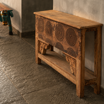 A rustic carved wooden console table with circular geometric engravings on the front, set against a textured gray wall and stone floor.