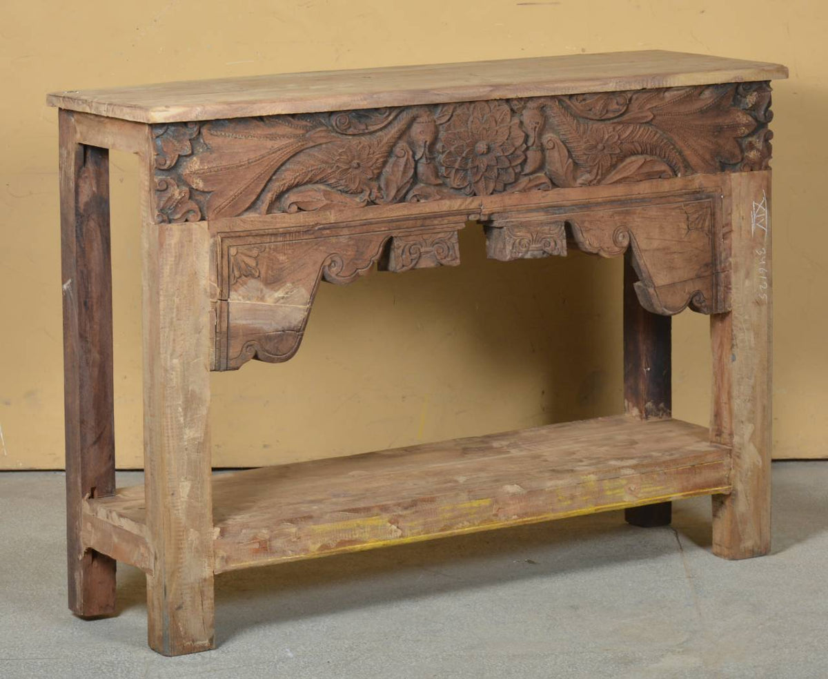 An antique carved wooden console table with a rustic finish, featuring intricate floral and scrollwork carvings on the front panel and a lower shelf, set against a plain yellow wall.