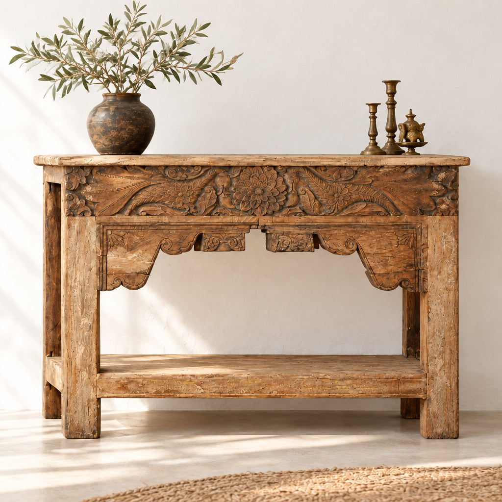A rustic carved wooden console table with a natural wood finish, featuring intricate floral and scrollwork carvings on the front panel and a lower shelf, set against a plain indoor wall.