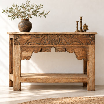 A rustic carved wooden console table with a natural wood finish, featuring intricate floral and scrollwork carvings on the front panel and a lower shelf, set against a plain indoor wall.