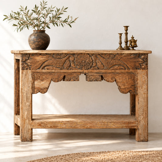 A rustic carved wooden console table with a natural wood finish, featuring intricate floral and scrollwork carvings on the front panel and a lower shelf, set against a plain indoor wall.