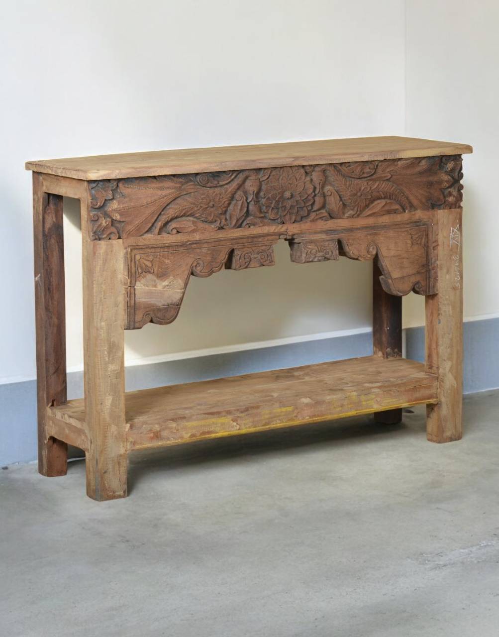 A rustic carved wooden console table with a natural wood finish, featuring intricate floral and scrollwork carvings on the front panel and a lower shelf, set against a plain indoor wall.