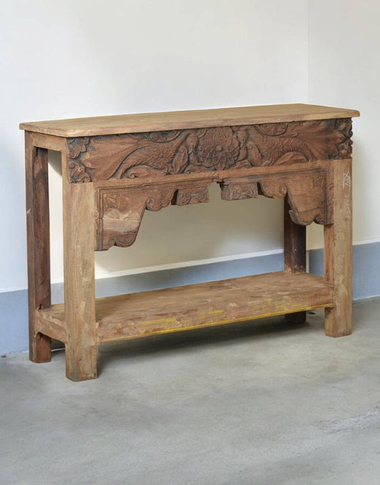 A rustic carved wooden console table with a natural wood finish, featuring intricate floral and scrollwork carvings on the front panel and a lower shelf, set against a plain indoor wall.