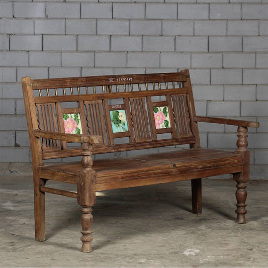 A rustic dark brown wooden bench with carved details and three colorful floral inlays on the backrest, set against a gray cinderblock wall.