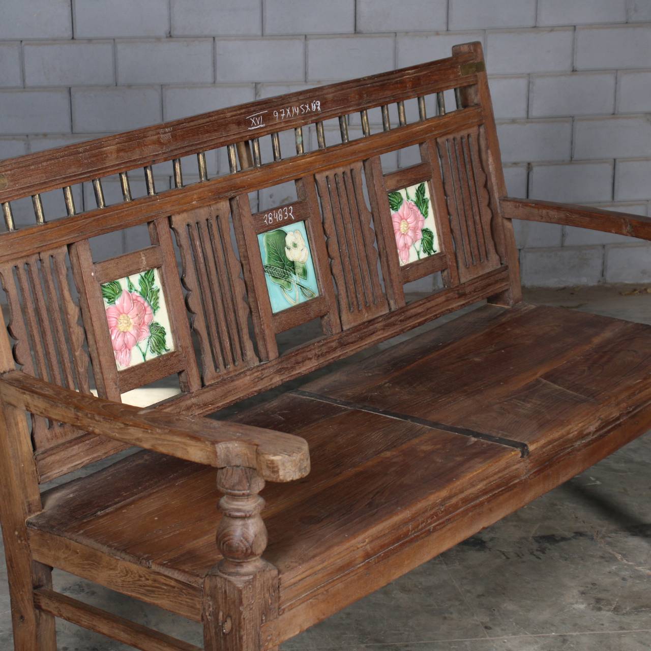 A rustic wooden bench with carved slats and three floral tile inserts on the backrest, photographed in a real interior setting with a gray brick wall and concrete floor.