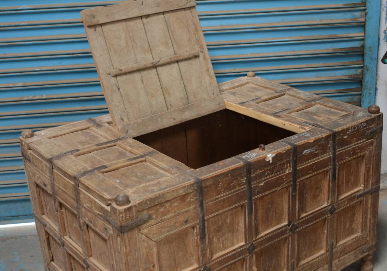 A large vintage wooden storage chest or trunk with an open hinged lid, showing a hollow interior. The chest has a paneled front and side, dark metal corner brackets, and brass-tone handles, photographed against a blue corrugated metal shutter.