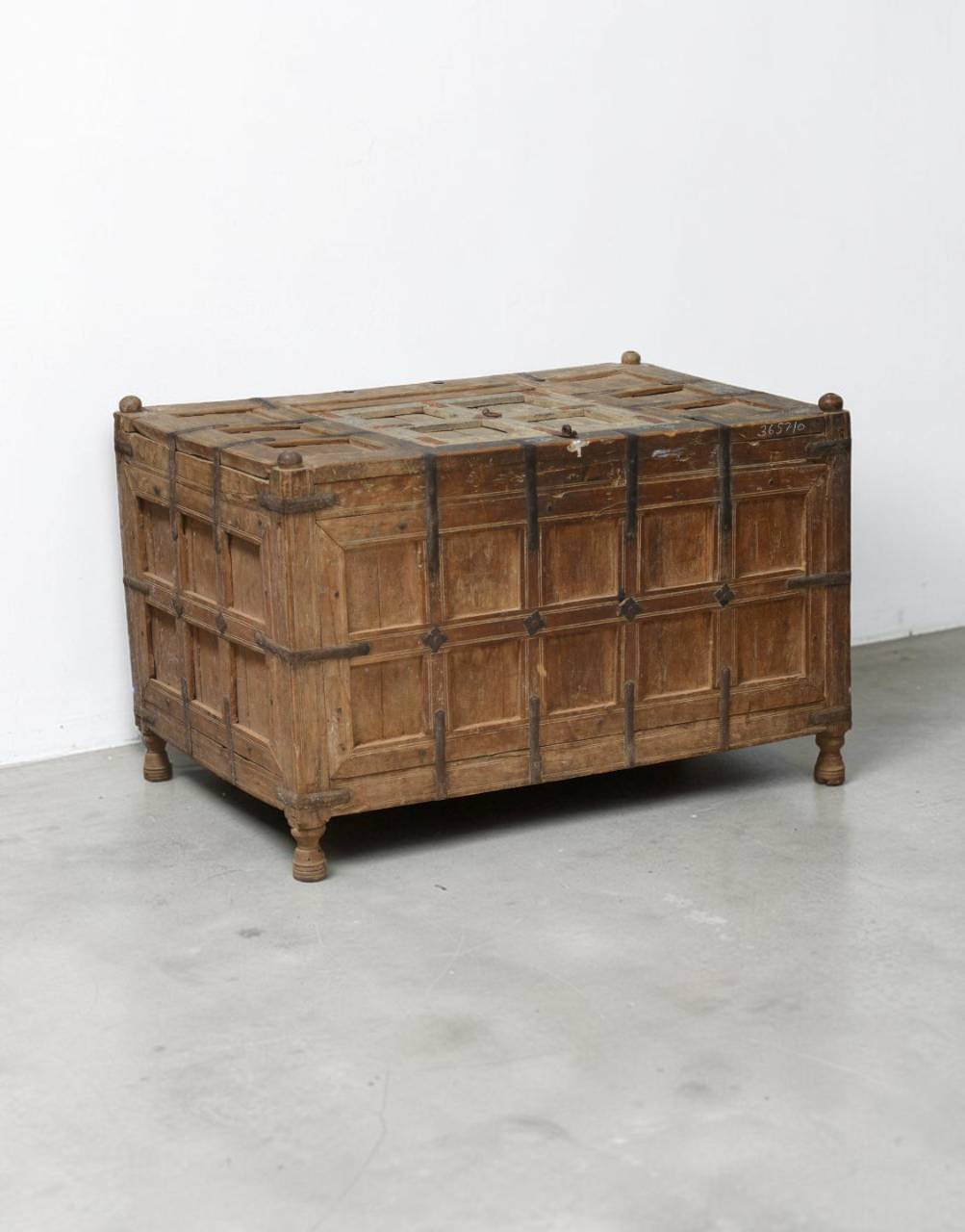 A rustic wooden chest with paneled front doors and dark metal straps, photographed in a minimalist studio setting with a white wall and gray floor.