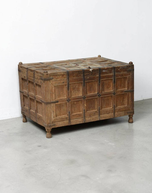 A rustic wooden chest with paneled front doors and dark metal straps, photographed in a minimalist studio setting with a white wall and gray floor.