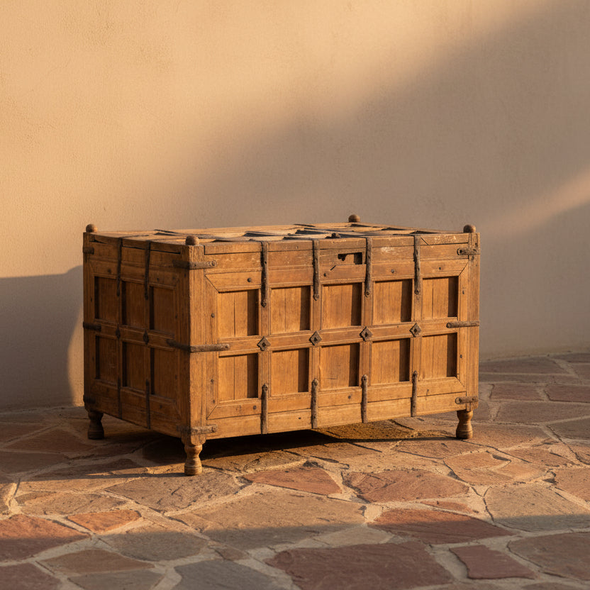 A rustic wooden chest with paneled front doors and dark metal straps, photographed in a minimalist studio setting with a white wall and gray floor.