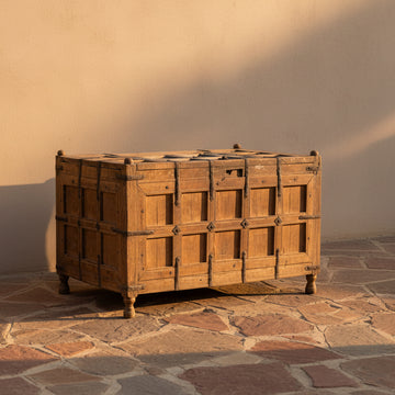 A rustic wooden chest with paneled front doors and dark metal straps, photographed in a minimalist studio setting with a white wall and gray floor.