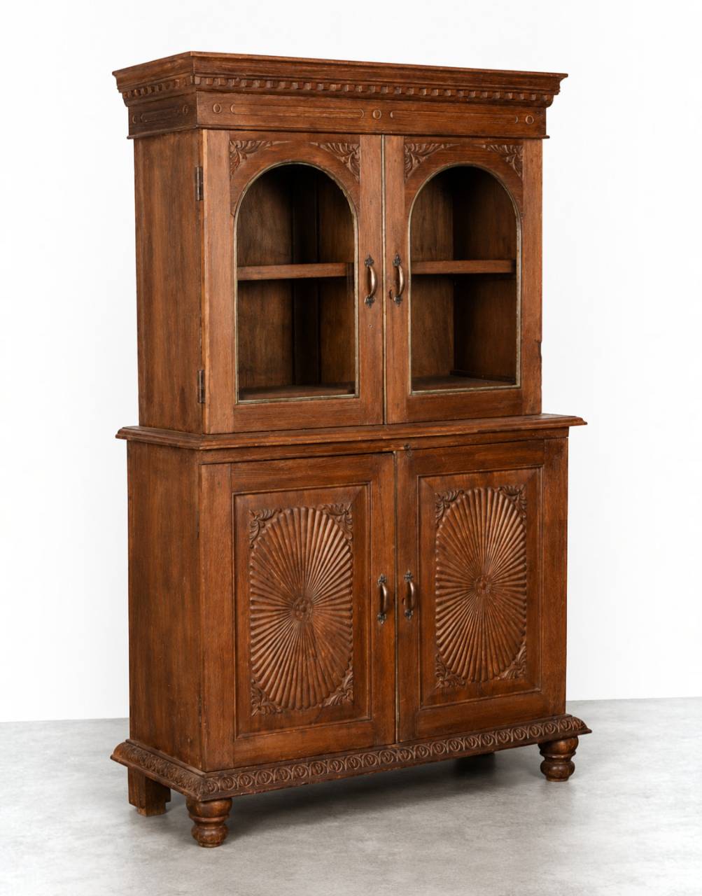 A tall, ornate wooden hutch cabinet with a warm medium-brown finish, featuring two upper arched glass-front compartments and two lower carved cabinet doors with sunburst detailing, set against a plain white studio background.