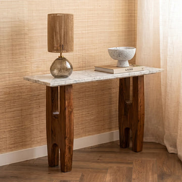 A rustic console table with a white marble top and dark brown carved wooden legs, styled with a lamp and a ceramic bowl in a warm, neutral-toned room.