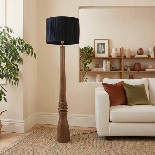 Floor lamp with a black shade and wooden base in a room with a plant and sofa.