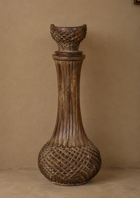 A tall, rustic carved wooden decorative vase with a fluted neck, textured diamond-patterned base, and a wide, scalloped rim, set against a warm beige stucco wall.