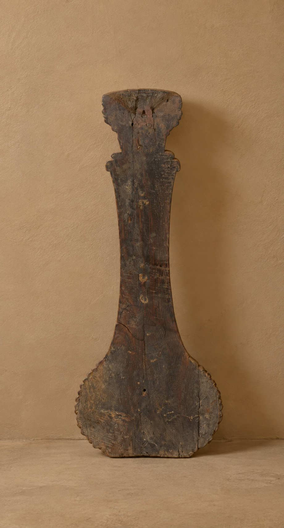 An antique dark brown wooden object with a weathered, rustic finish, standing upright against a textured beige plaster wall.