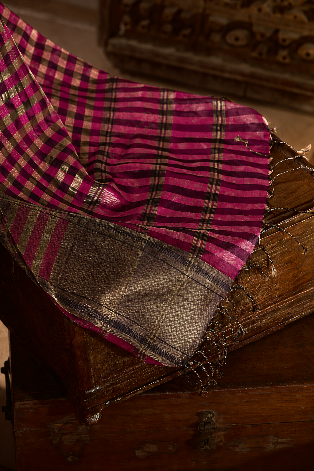 A close-up of a draped pink and black checkered fabric, likely a sari or scarf, with a metallic gold border, resting on an antique wooden chest.