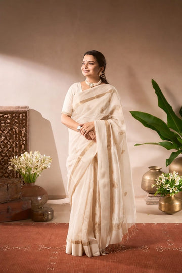 A woman wearing a light beige/cream saree with gold border details, standing in a styled indoor setting with decorative plants, vases, and a textured wall.
