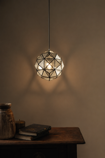 A geometric faceted pendant light with a warm glowing bulb inside, hanging above a rustic wooden table with books and a ceramic jar against a dark brown wall.