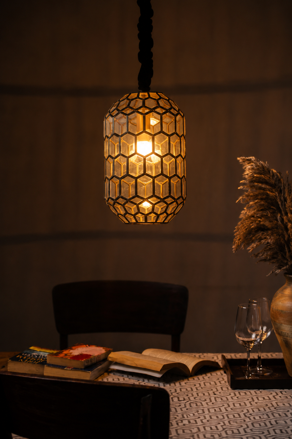 A warm glowing pendant light with a honeycomb patterned shade hangs over a dining table in a dimly lit room. Books, a wine glass, and dried pampas grass are visible on the table.