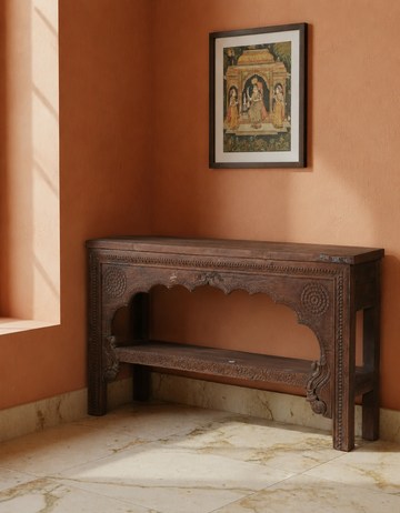 An ornate dark brown carved wooden console table placed in a warm terracotta-colored room corner with a framed artwork hanging on the wall above it.