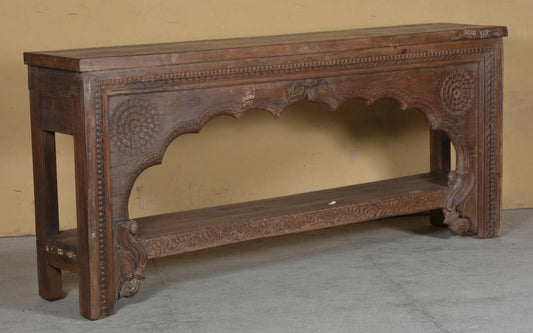 An ornate dark brown carved wooden console table with intricate floral and geometric patterns, set against a plain beige wall and gray floor.