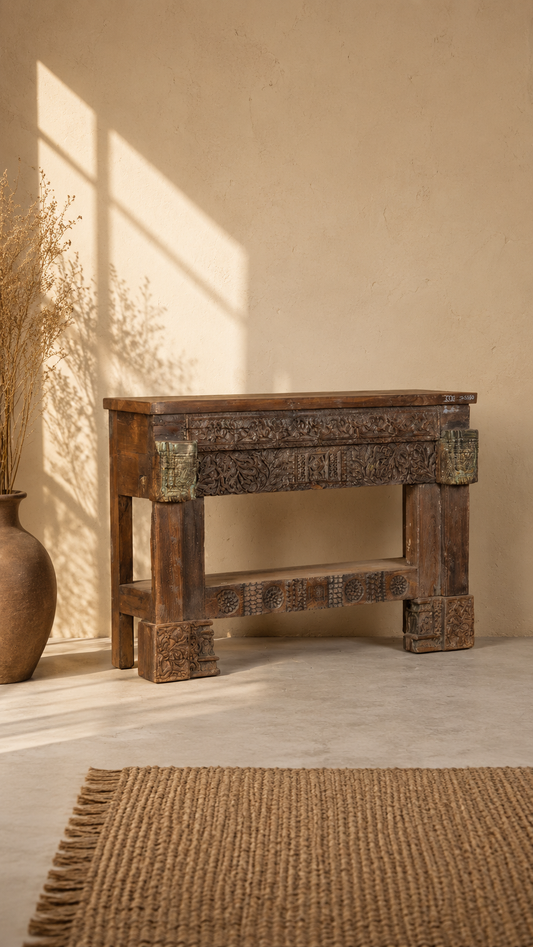 An antique-style carved wooden console table with intricate relief carvings, set in a warm, sunlit room with a textured beige wall.