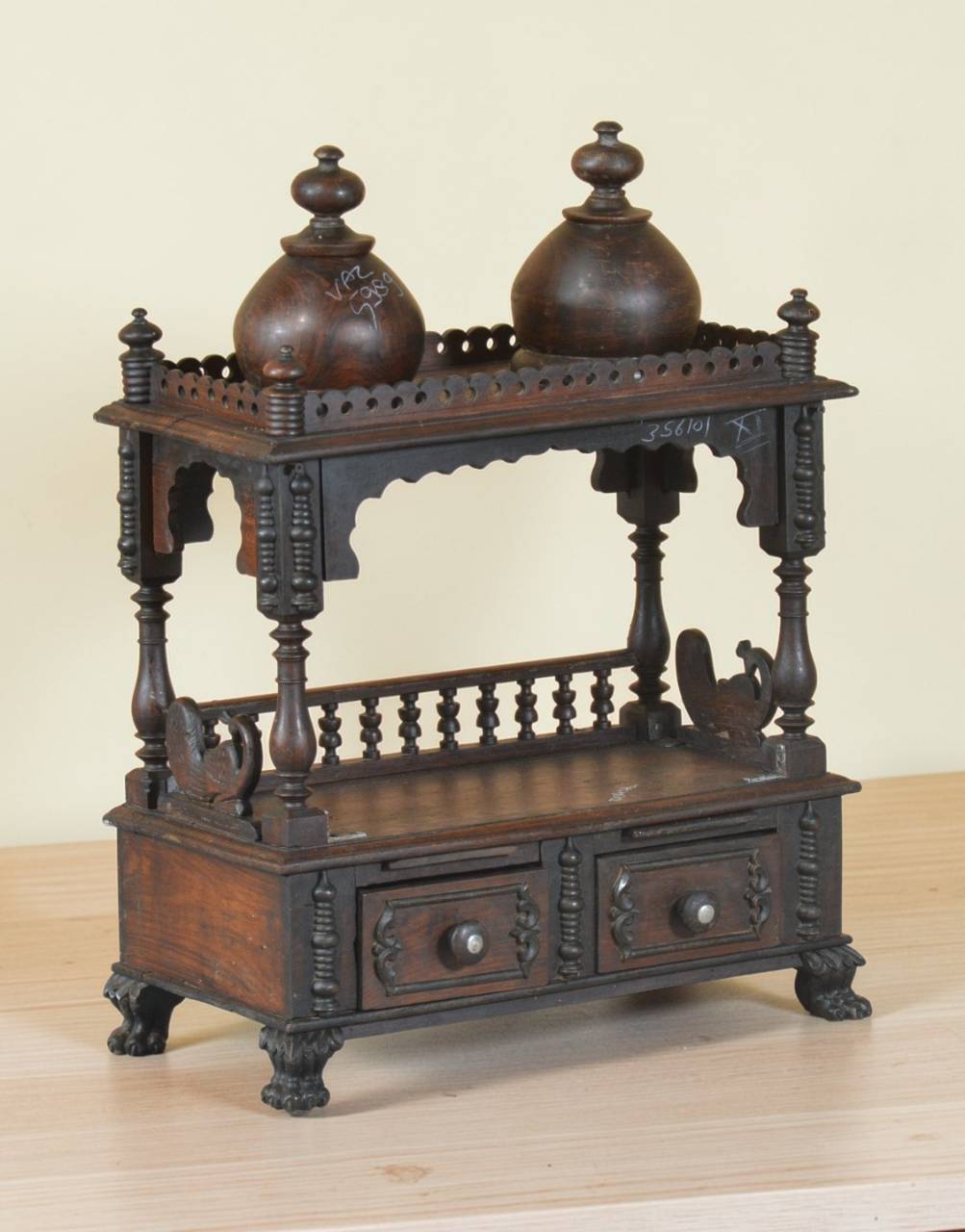 An ornate dark brown carved wooden miniature cabinet or display stand with two small decorative lidded vessels on top, photographed against a plain light beige wall and pale wood surface.