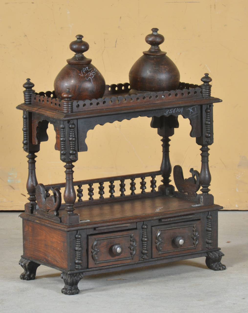 An antique dark brown carved wooden cabinet with two drawers, a decorative upper shelf, and two large matching wooden bottles on top, set against a plain beige wall.
