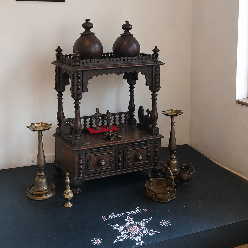 A dark carved wooden deity stand or small altar with two dome-shaped vessels on top, displayed indoors on a raised dark platform against a light wall. The scene includes hanging black lanterns, framed wall art, brass candle holders, and a white decorative motif on the platform floor.