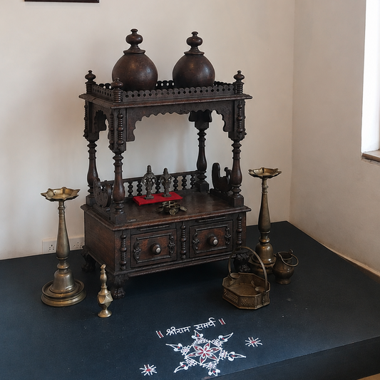A dark carved wooden deity stand or small altar with two dome-shaped vessels on top, displayed indoors on a raised dark platform against a light wall. The scene includes hanging black lanterns, framed wall art, brass candle holders, and a white decorative motif on the platform floor.