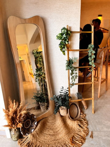 A large floor mirror with a wavy light wood frame leans against a textured wall in a warm, boho-style room. The scene is decorated with a wooden ladder shelf draped with green trailing plants, a round woven rug, dried pampas grass in a glass vase, and a straw hat resting on the floor.