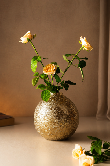 A round, textured metallic gold vase holding three pale yellow roses with green stems, set against a warm beige background.