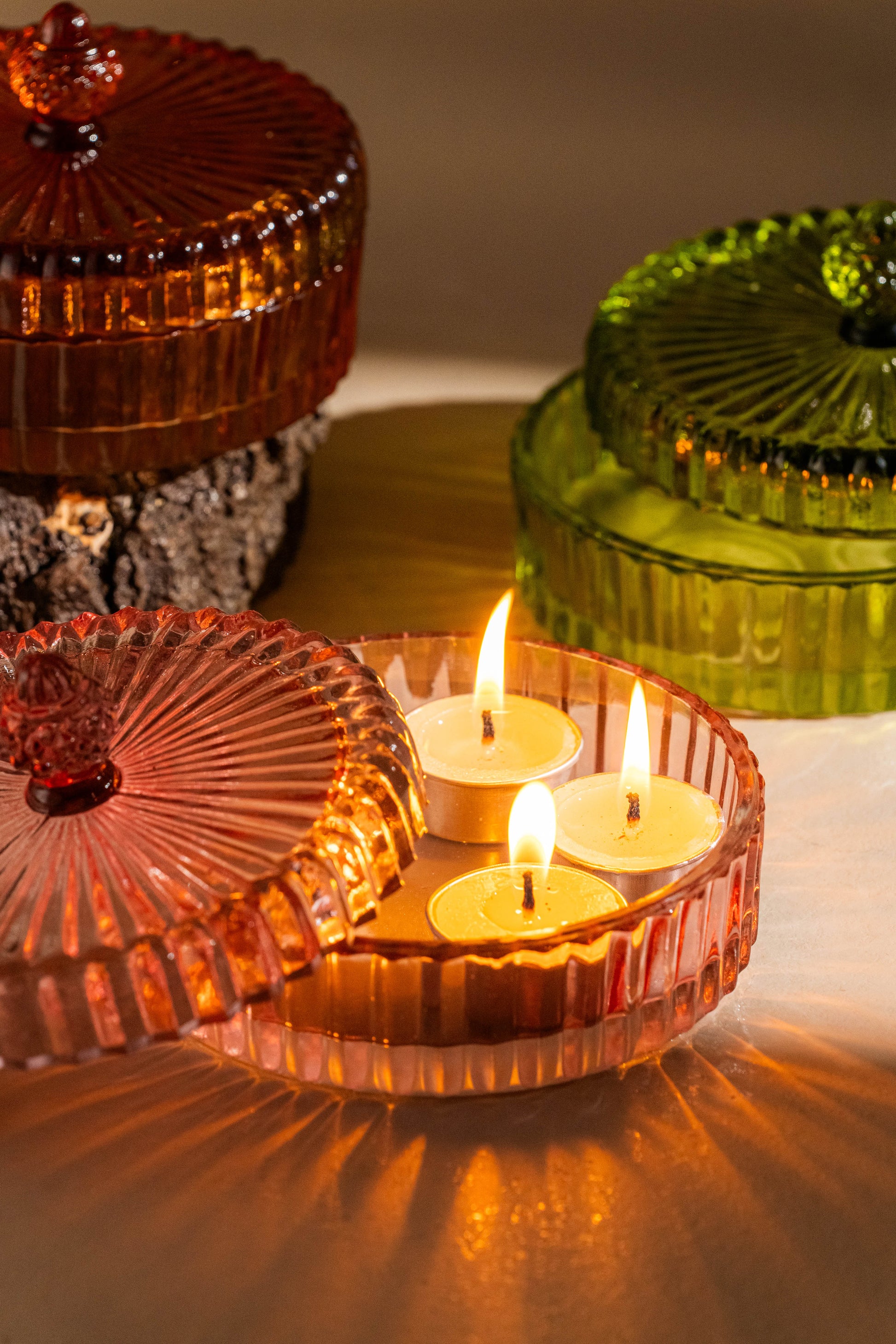 Three lit candles in a decorative glass holder with colorful lids on a soft surface.