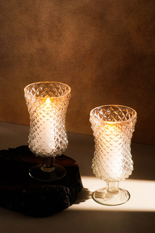 Two textured glass candle holders with lit candles on a dark surface and brown background