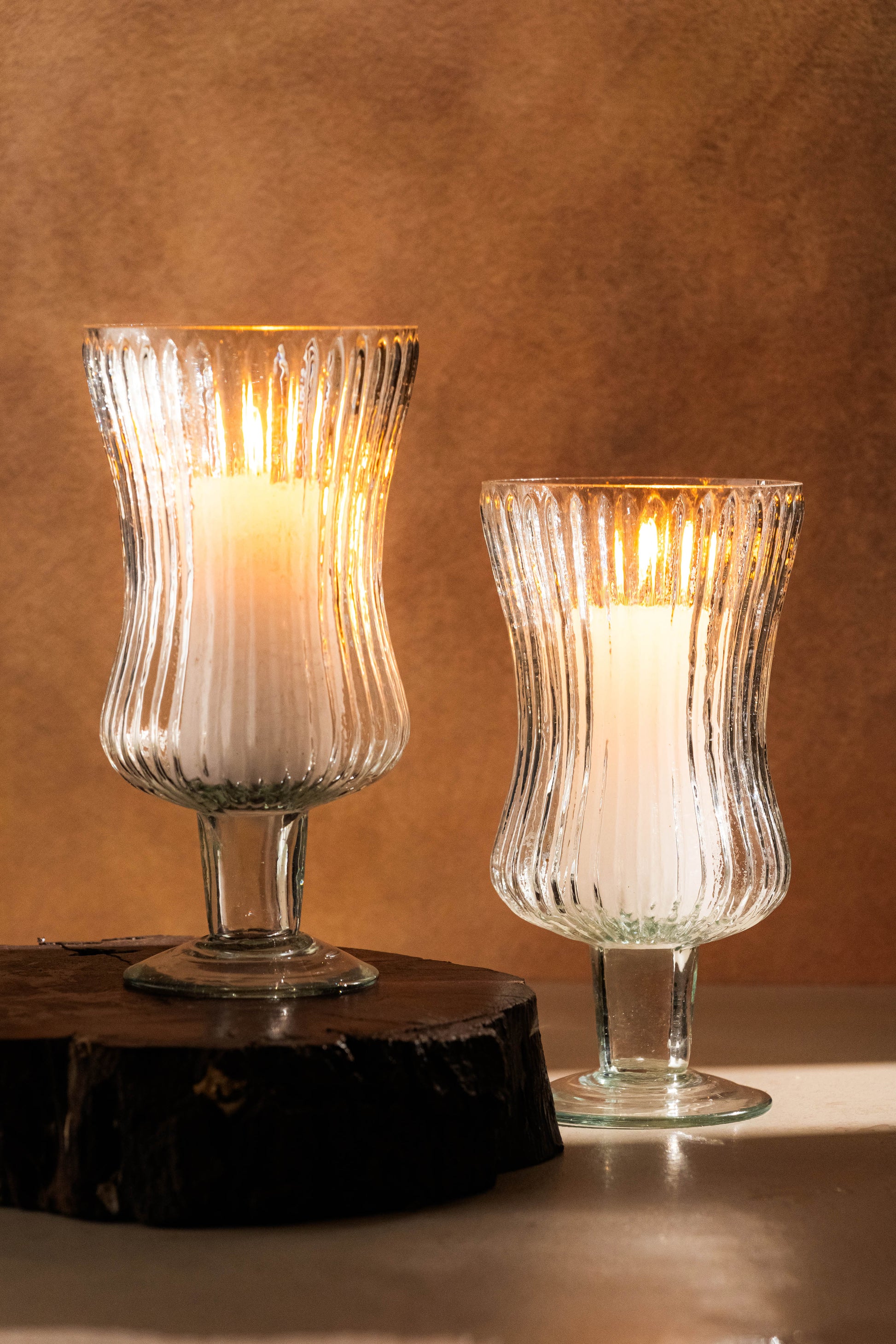 Two glass candle holders with lit candles on a wooden surface against a brown background