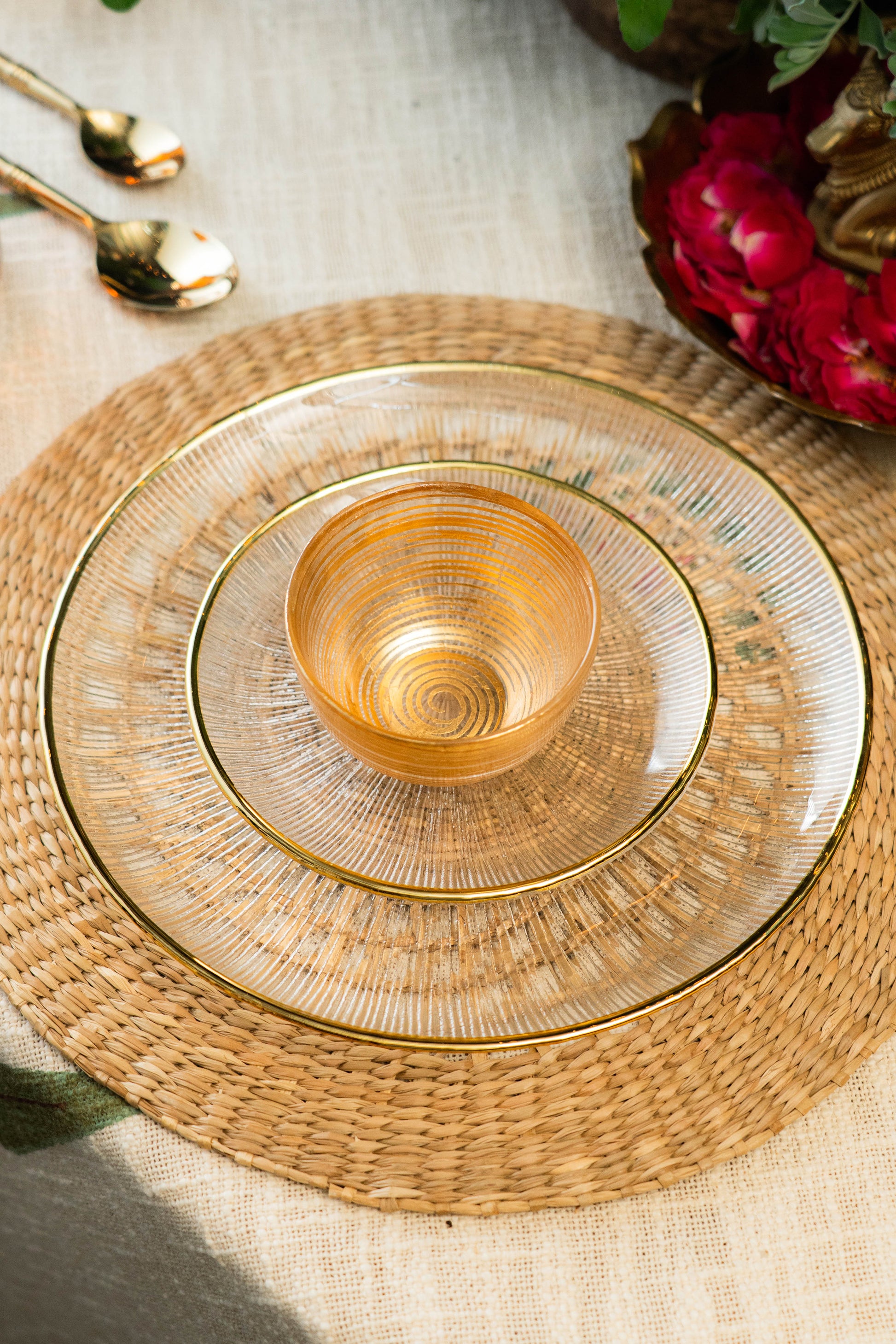 Gold glass plate with a woven placemat on a textured surface with flowers and cutlery.