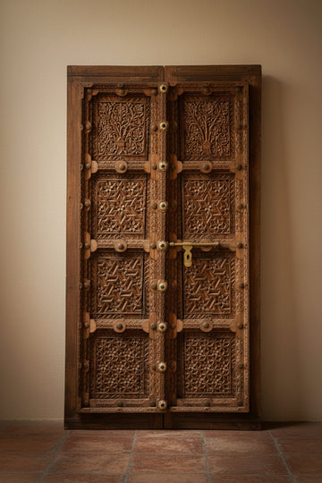 A pair of tall, intricately carved dark brown wooden doors with brass hardware, set against a plain white wall and light gray floor.