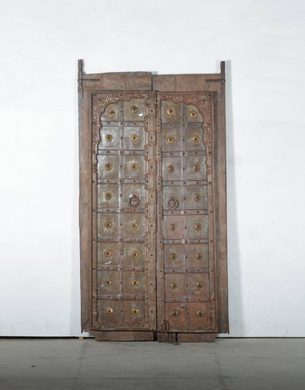 A pair of tall, rustic wooden doors with carved details and numerous round brass-toned metal studs, standing against a plain white wall.