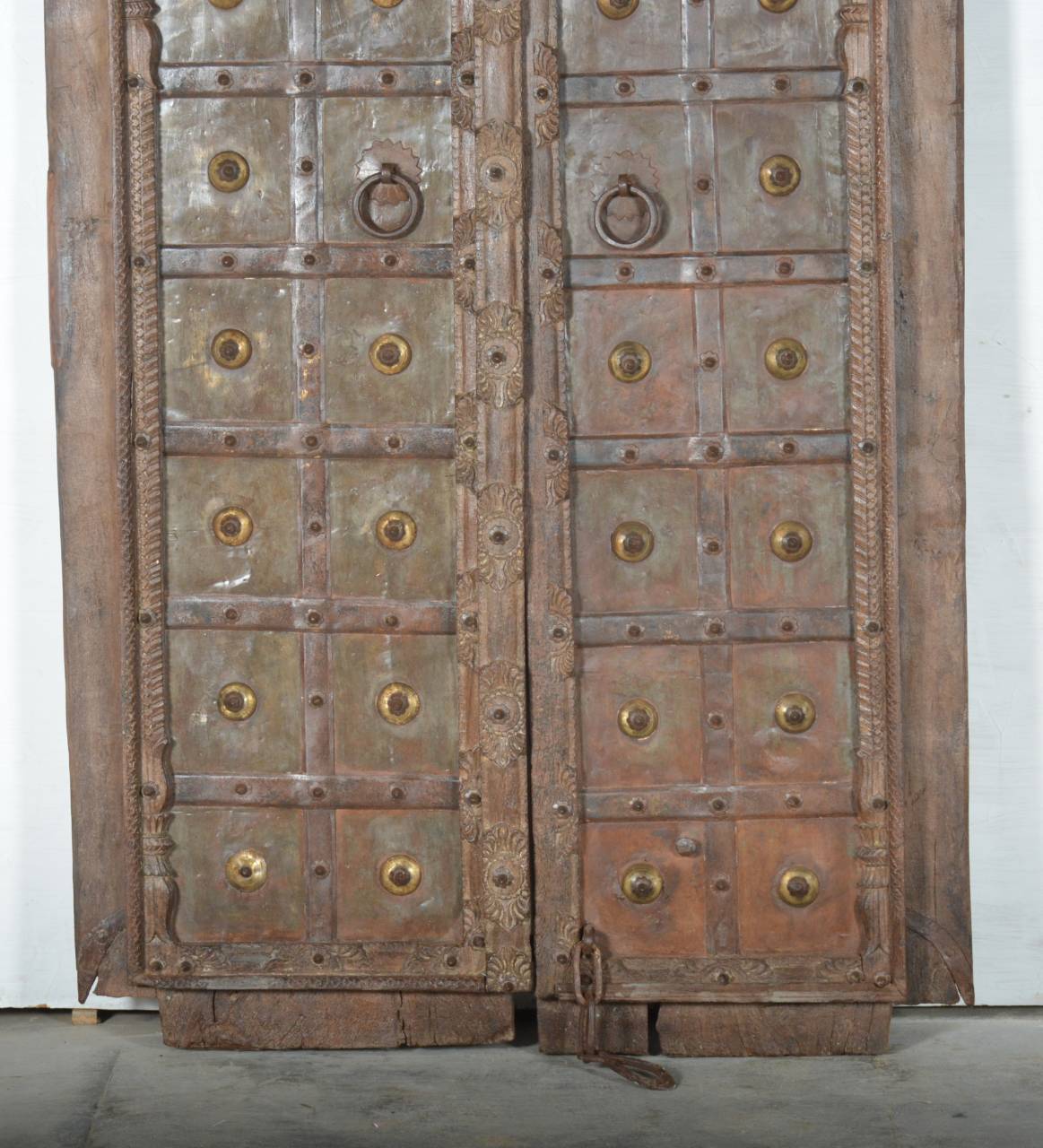 An antique double wooden door with heavy metal plates, brass studs, and circular door knobs, photographed against a plain white wall.